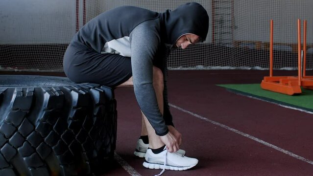 Sportsman tying shoelace while sitting on heavy tire. Male athlete is preparing before workout at gym. He is wearing hooded shirt during training.