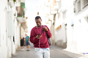 Smiling young black man with mobile phone listening to music with earphones in the city