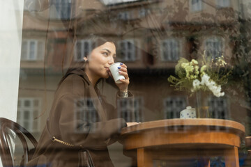 young woman in autumn coat drinking coffee near window in prague cafe.