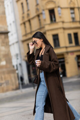 Obraz premium Side view of stylish tourist in coat and sunglasses holding takeaway coffee on street in Prague.