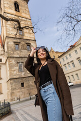 Fototapeta premium Low angle view of happy traveler in coat holding sunglasses near blurred buildings in Prague.