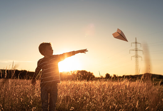 Little child flying his paper airplane up in the sky. Dreams of traveling in summer in nature at sunset
