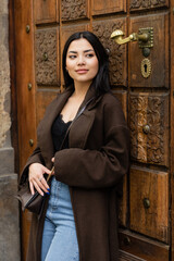 smiling woman in trendy coat with crossbody leaning on carved door and looking away in prague.