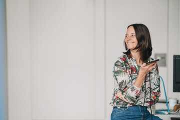 Business woman sitting on desk in office with cellphone and looking happily to the side