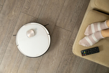 White robotic vacuum cleaner cleans laminated floor in light living room. Female feet in striped...