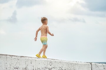 Preschooler boy in bright colorful shorts walks on sea concrete pier against cloudy sky. Child enjoys summer holidays on sea