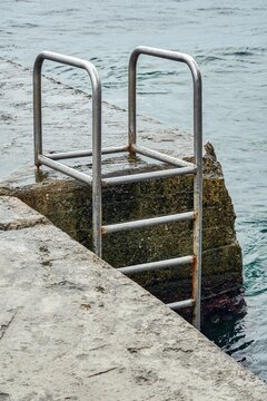 Grey concrete pier with metal ladder at blue sea water with small waves on an overcast summer day