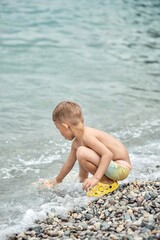 Preschooler boy plays on pebble beach of sea catching light waves of water. Excited child sticks out tongue enjoying playing near sea water edge