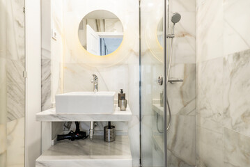 Bathroom tiled in white marble with gray veins, circular mirror and glass shower cubicle with square sink on marble shelves