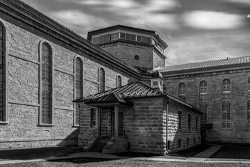 Exterior view of two large limestone penitentiary cell blocks meeting at a central guard post, high barred windows, high key black and white, nobody