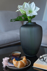 Cup of coffee, croissant, book with glasses and flowers  on round coffee table. Fashionable interior as background.