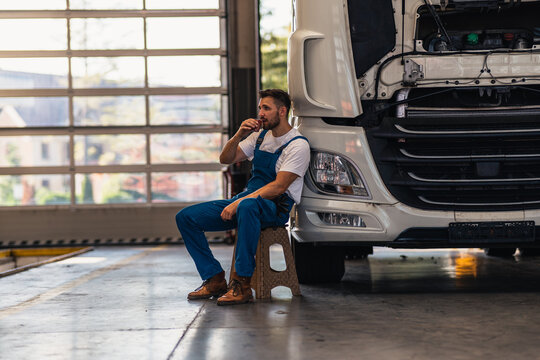 A bottom view of a mechanic drinking tea or coffee from a cup and sitting leaning against a truck in a workshop. A young caucasian men mechanic. Truck service, repair. Rest and break at work.