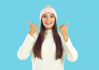 Portrait of happy friendly girl with long hair in light pink hat and white sweater showing thumbs...