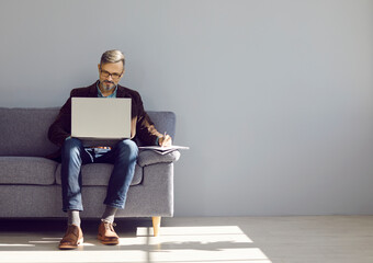 Modern businessman working on laptop. Mature man in stylish smart casual clothes working on notebook computer and writing down information while sitting on sofa near grey copy space wall