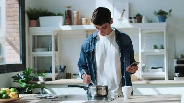 Video of handsome young man cooking and using his mobile phone in the kitchen at home.