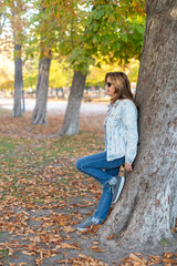 Mature white woman leaning against a large tree in a public park with leaves fallen on the ground by autumn.