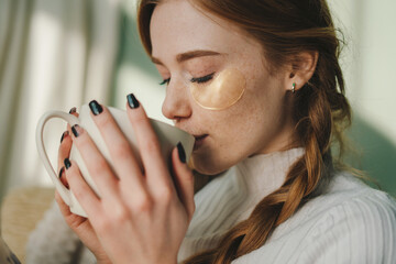 Portrait of a beautiful freckled woman with eye-patches resting in cozy sofa drinking a cup of tea, enjoying weekend time. Under eye patches. Leisure, rest