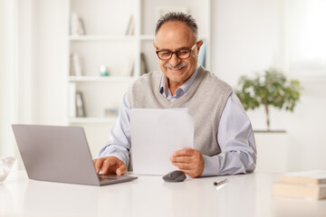 Smiling mature man holding a paper document and sitting in front of a laptop computer
