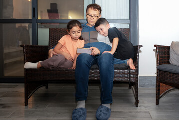 Grandmother and her grandchildren use cell phone together while sitting on couch on terrace.