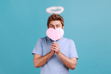 Portrait of angelic man with holy nimbus covering half of face with pink paper heart on stick,...