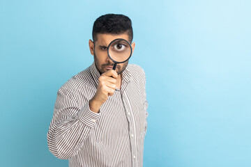 Serious bearded man holding magnifying glass and looking at camera with big zoom eye, having strict...