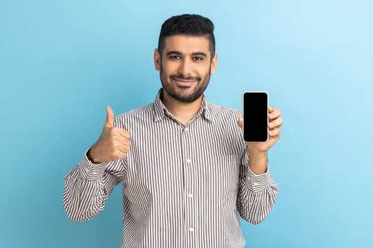 Young satisfied businessman standing holding phone with empty display, showing thumbs up with toothy smile, looking at camera, wearing striped shirt. Indoor studio shot isolated on blue background.