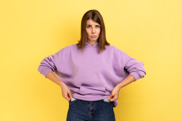 Unhappy poor woman turning empty pockets inside out and looking frustrated by overspend, lack of money, financial crisis, wearing purple hoodie. Indoor studio shot isolated on yellow background.