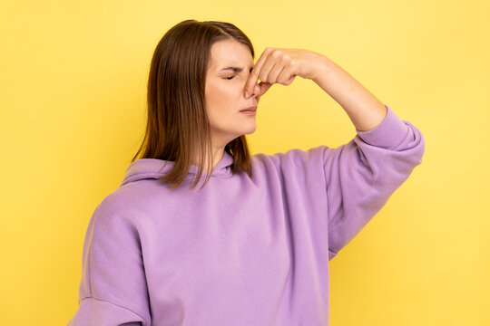 Portrait of dark haired woman grimacing with disgust, holding breath, pinching nose with fingers to avoid bad smell, awful odor, wearing purple hoodie. Indoor studio shot isolated on yellow background