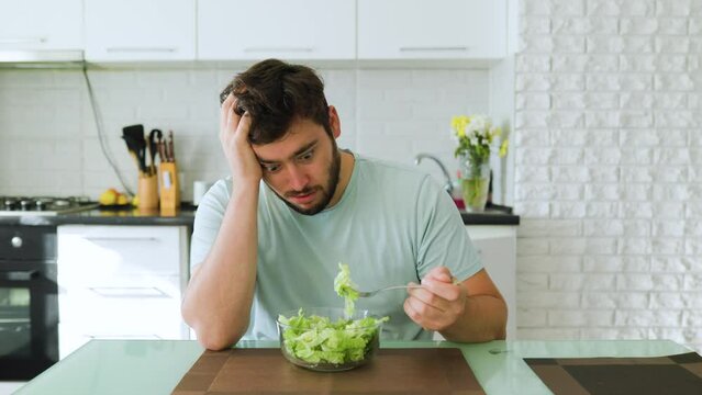 A sad young man looks longingly at a salad, holds a fork with greens in his hand, and a plate of salad in front of him. An exhausting diet haunts, hates healthy food. Stop diet.