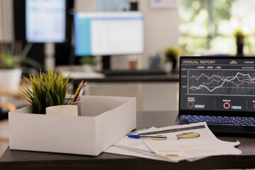 Sheets with printed statistical reports placed on desk in modern financial with no people on it. Empty workstation with plant and coffee cup in cardboard box, annual statistic concept.
