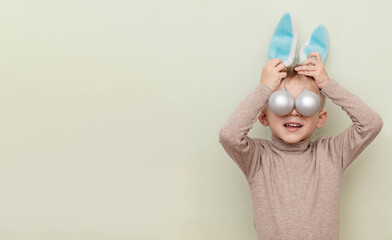 A funny boy in the guise of a rabbit holds two silver matte toys, covering his eyes with them on a light background with a space for copying. The concept of the New Year