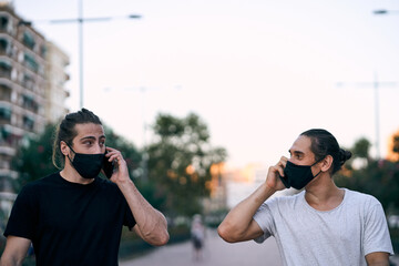 two caucasian young men walking on the city boulevard wearing face mask talking on the smartphone looking at each other