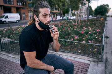 young caucasian man in black t-shirt and blue pants sitting vaping with a black respirator under his mouth in the city