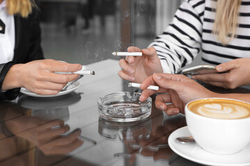 Women holding cigarettes over glass ashtray at table, closeup