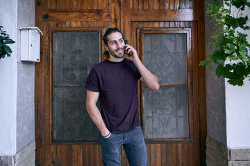 caucasian young man with beard talking on the smartphone of him smiling near the wooden door and mailbox