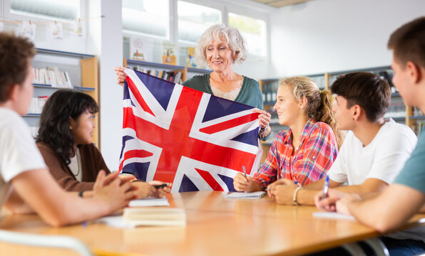 Elderly teacher shows the flag of Great Britain to teenagers and talks about this country during lesson at a library school