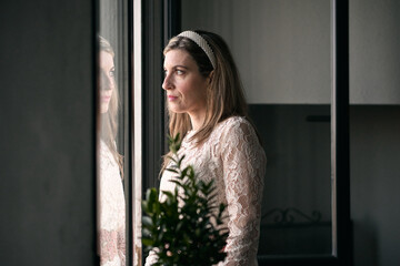 Fototapeta na wymiar caucasian mature woman in white dress and headband in her hair standing looking out the window calm and relaxed next to a plant