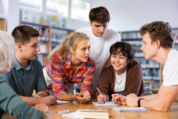 Group of positive young teen students communicating in school library, talking and making notes