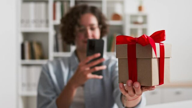 A girl takes a picture of a gift box with a red bow, a blogger shoots a gift with a red ribbon on a camera, a gift is filmed on a smartphone smartphone
