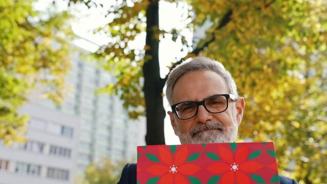 Middle-aged man with grey hair and beard sitting on park bench holding red gft bag smiling to camera. Birthday gift. Horizontal outdoor video. High quality 4k footage