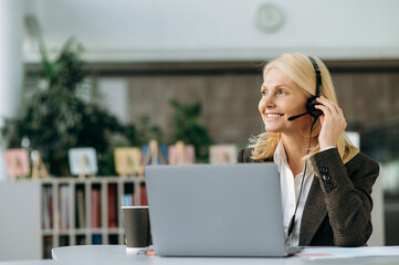Elegant happy business woman is working as operator of call center, sitting at the desk in office. Mature female employee in headset and stylish suit consulting clients, smiling