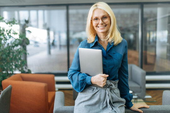 Portrait of happy confident businesswoman in eyaglasses and stylish formal suit. Middle aged successful female leader looking directly at the camera, smiling, holding laptop in arms