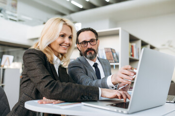 Fototapeta na wymiar Male and female employee in formal suits are sitting at the work desk in office. Smart senior man consulting beautiful woman about work process. Concentrated colleagues using laptop, working together