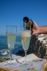 Pouring of champagne sparkling wine into glasses with view on white houses of picturesque village Azenhas do mar, Lisbon area, Portugal