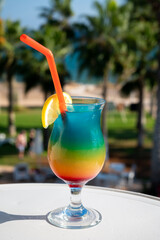 Colourful cold Rainbow Paradise cocktail drink served in glass at pool bar overlooking blue pool, sea and palm trees, relax and holidays at sea