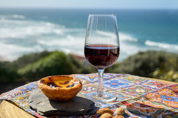 Portugal's traditional food and drink, glass of porto wine and sweet dessert Pastel de nata egg custard tart pastry served with view on blue Atlantic ocean in Lisbon area, Portugal - obrazy, fototapety, plakaty