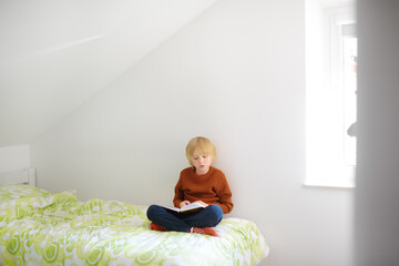 Smart preteen boy sitting on bed and enthusiastically reading interesting book. Kid reader enjoying...