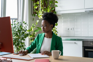 Focused woman freelancer works remotely performing tasks of large companies via Internet. Thoughtful African American girl in green business jacket sits at table with computer and cup of coffee 