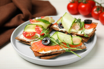 Tasty rye crispbreads with salmon, cream cheese and vegetables served on white wooden table, closeup