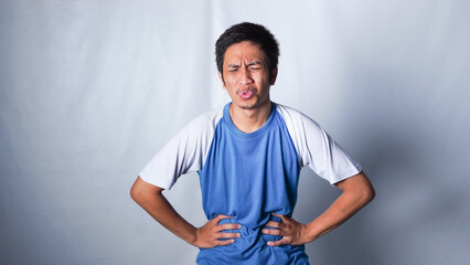 Young asian man wearing blue shirt standing over isolated white background touching stomach with hand with painful expression because of stomachache